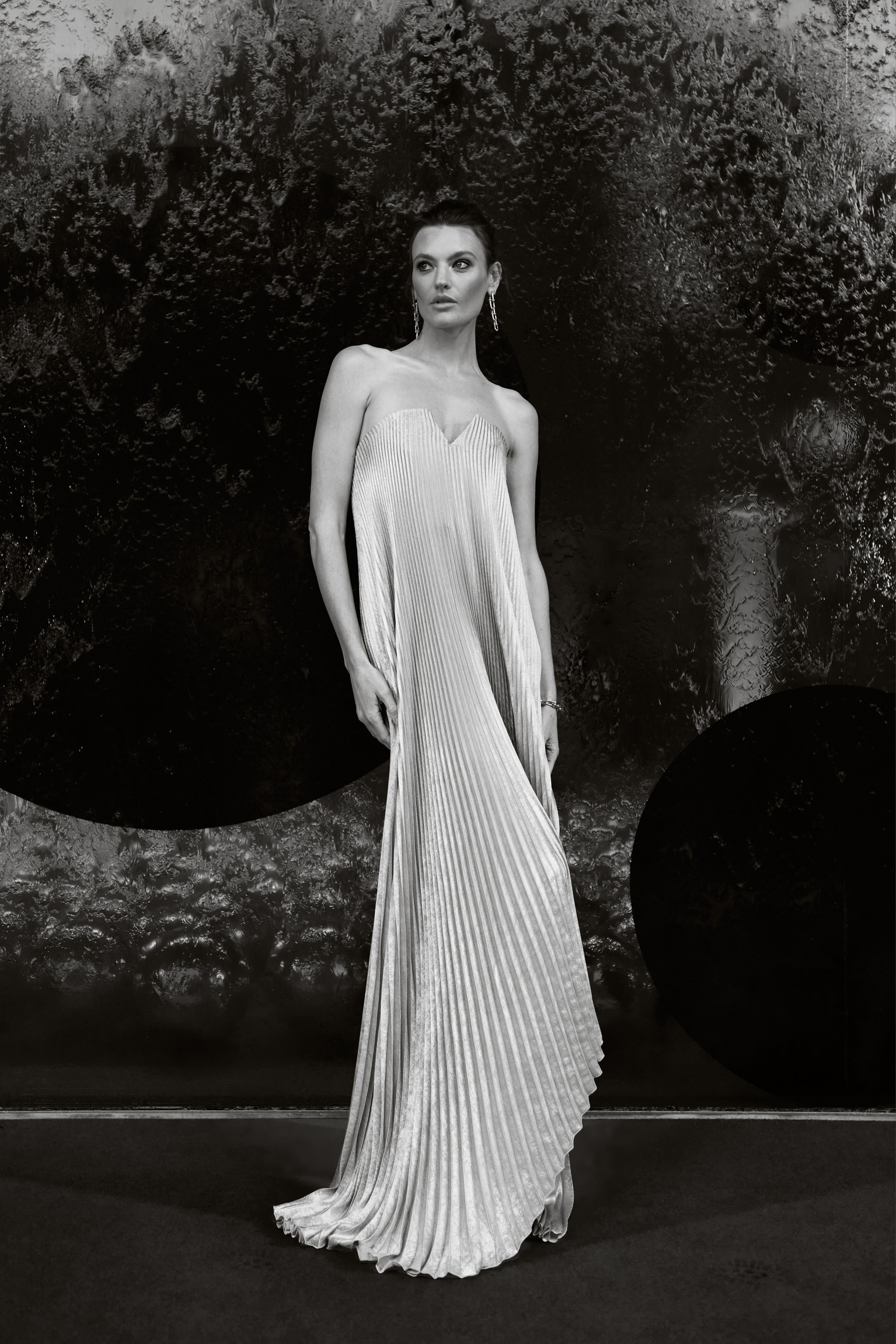 Elegant adult female in a silver gown posing in a studio with a textured black backdrop, wearing jewelry and styled hair.